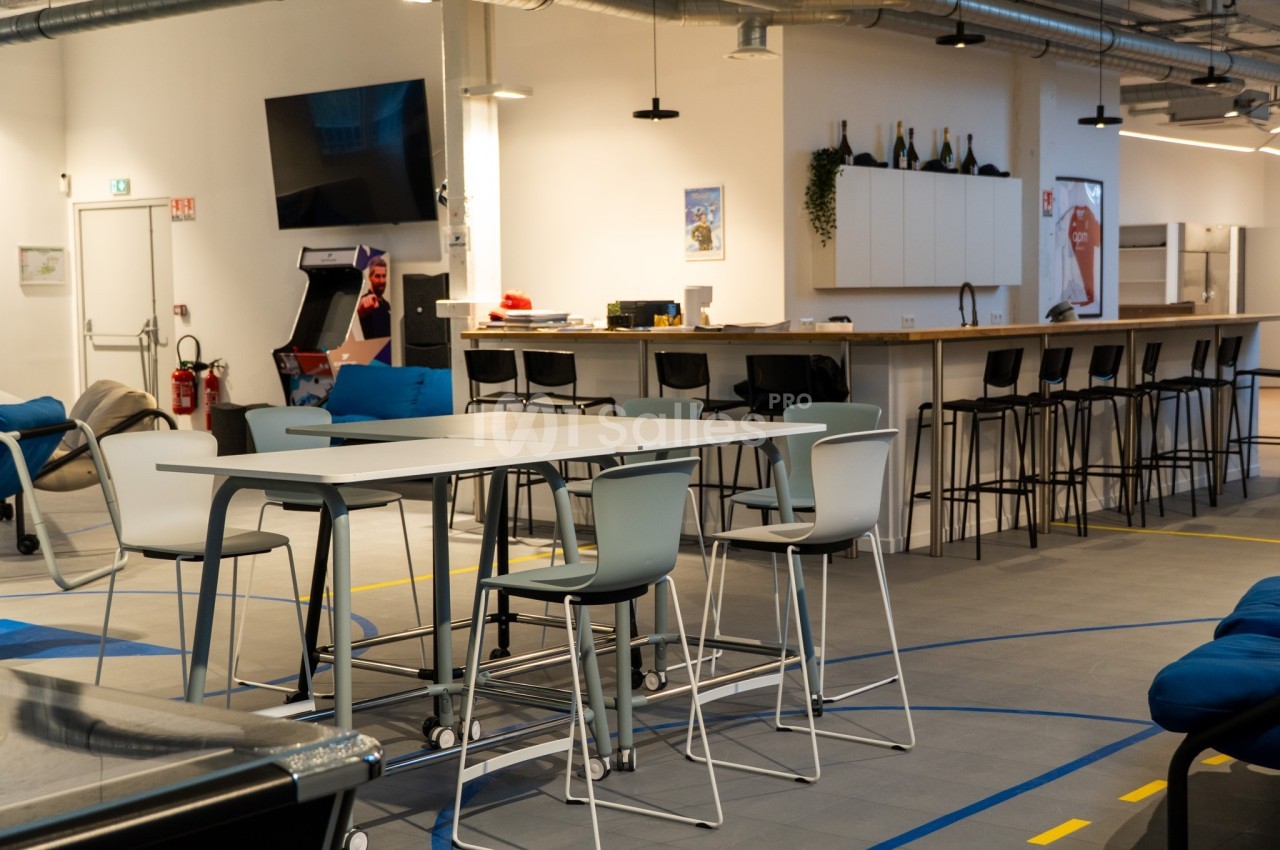 Espace de détente moderne avec tables hautes, chaises, bar, canapés et décor industriel lumineux.