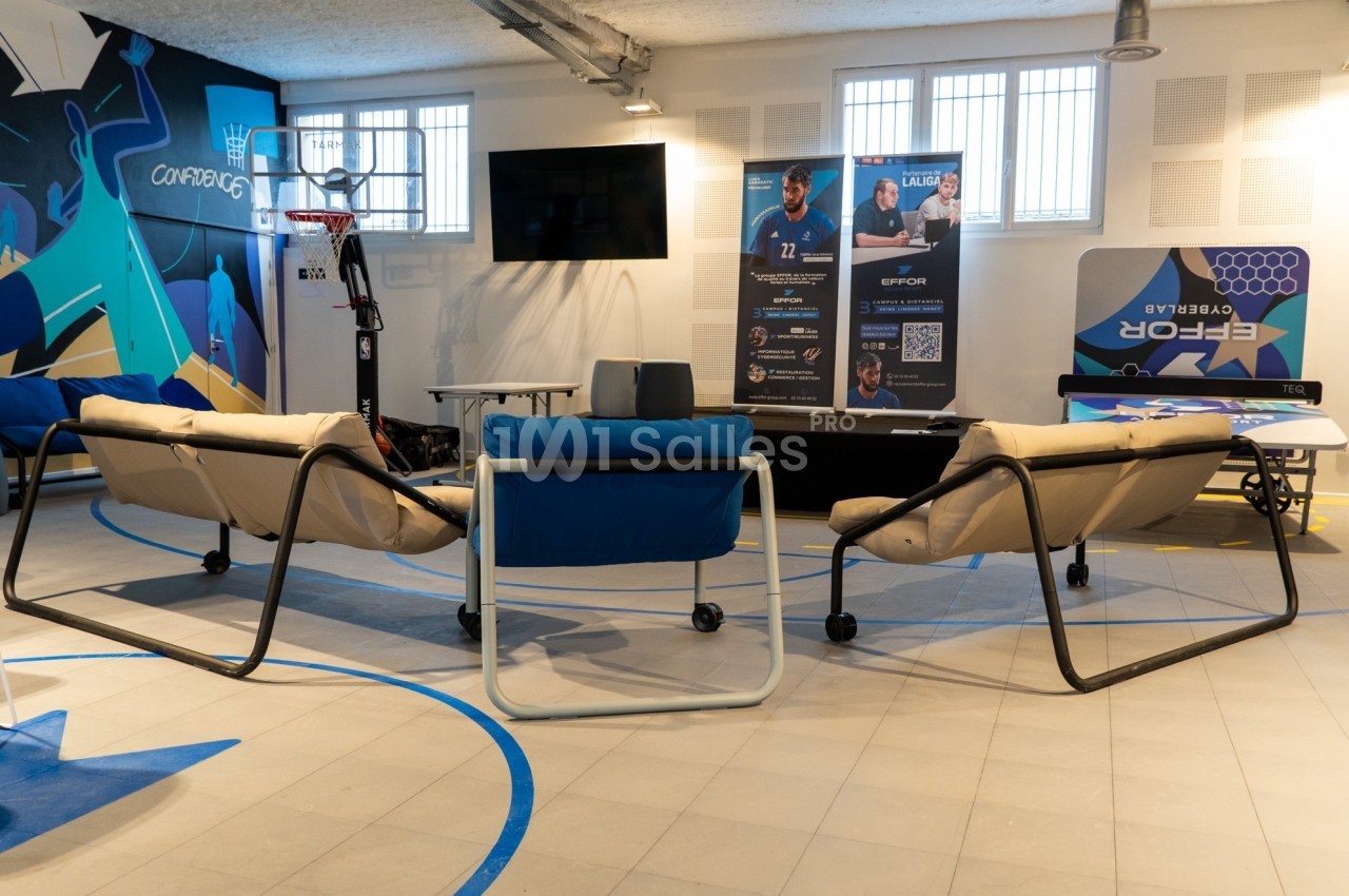 Espace intérieur avec canapés, écran mural, affiches promotionnelles et équipements sportifs visibles en arrière-plan.