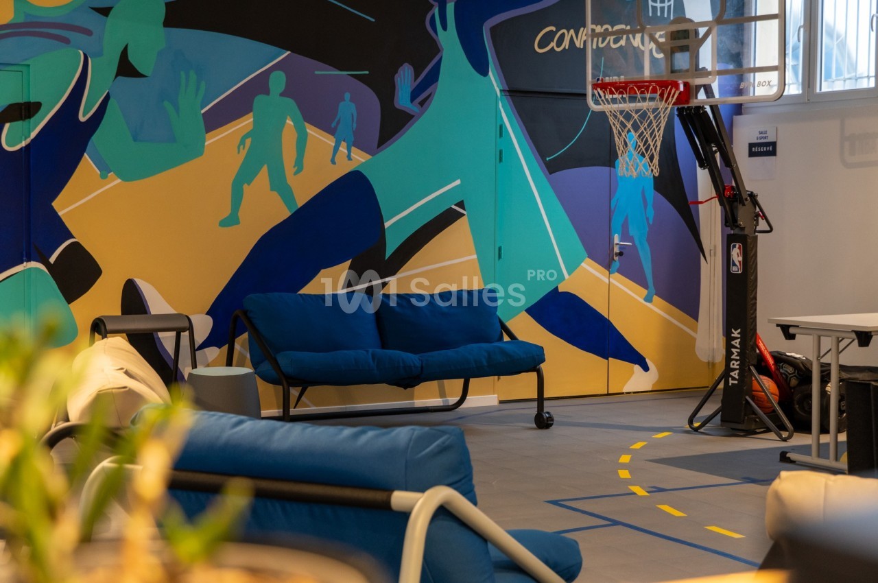 Mur coloré avec des silhouettes de joueurs de basket, canapé bleu et panier de basketball dans un espace intérieur lumineux.