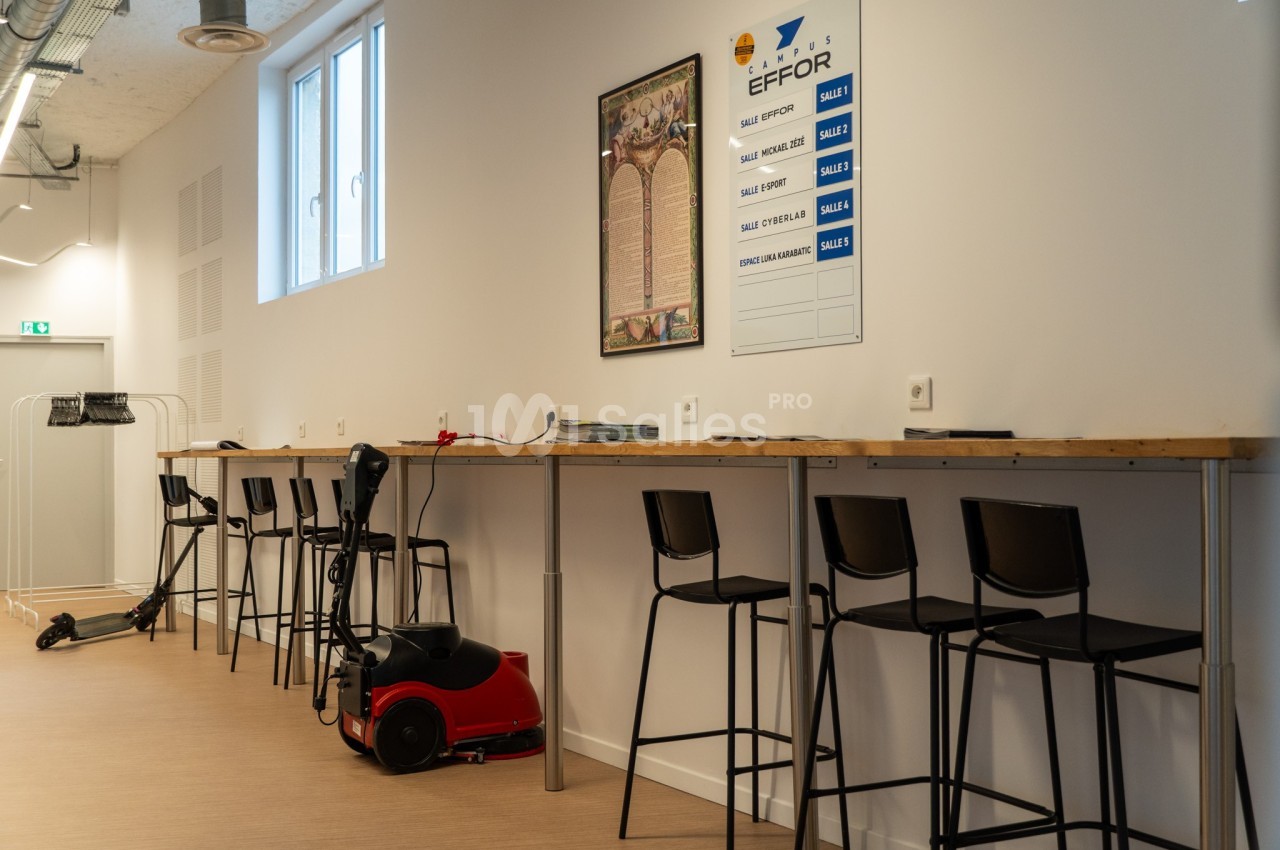 Salle équipée de tabourets hauts alignés le long d'un comptoir, avec un aspirateur et un tableau mural en arrière-plan.