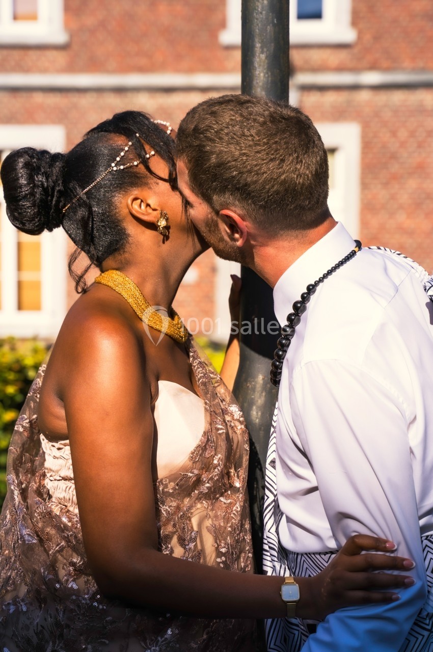 Un couple s'embrasse en extérieur devant un bâtiment en briques, sous une lumière ensoleillée.
