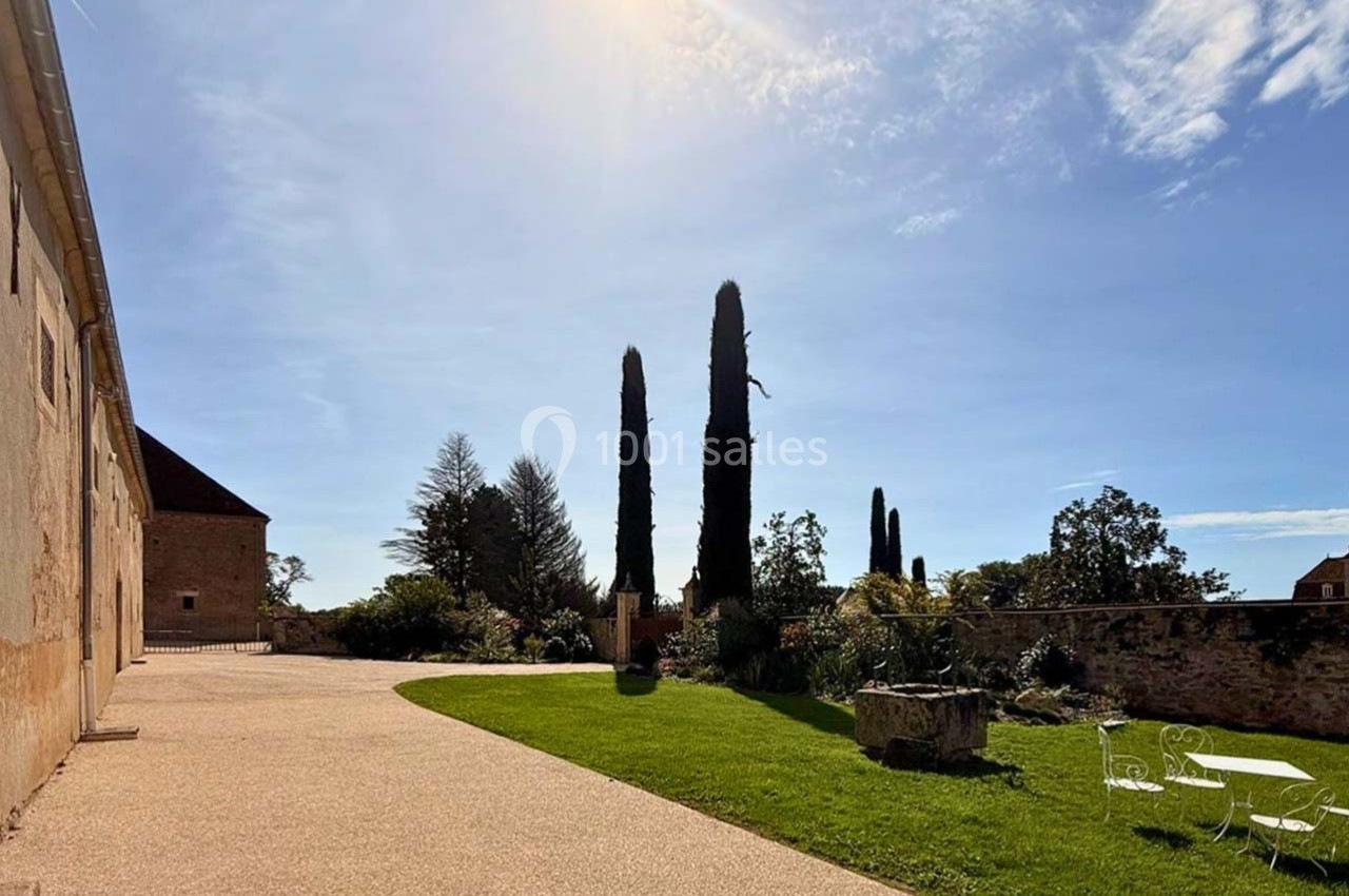 Cour ensoleillée avec allée pavée, pelouse, cyprès et mobilier de jardin, entourée de bâtiments en pierre.