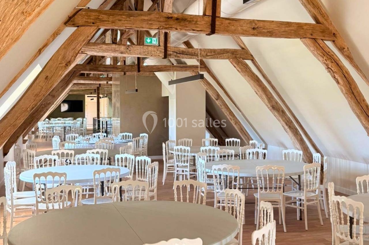 Salle lumineuse sous charpente en bois avec des tables rondes et des chaises blanches disposées pour un événement.