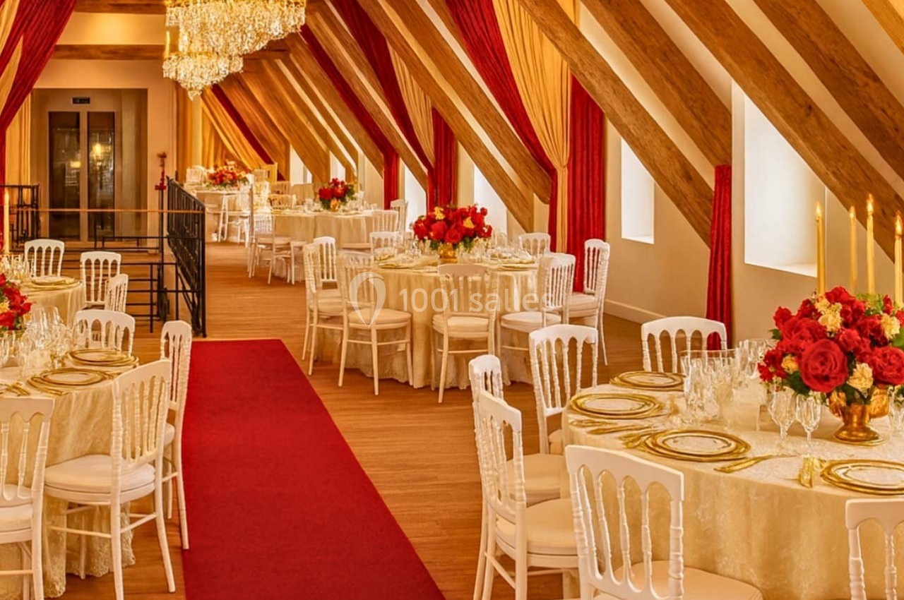 Salle de réception élégante avec tables décorées, nappes blanches, chandeliers dorés, fleurs rouges et tapis rouge.