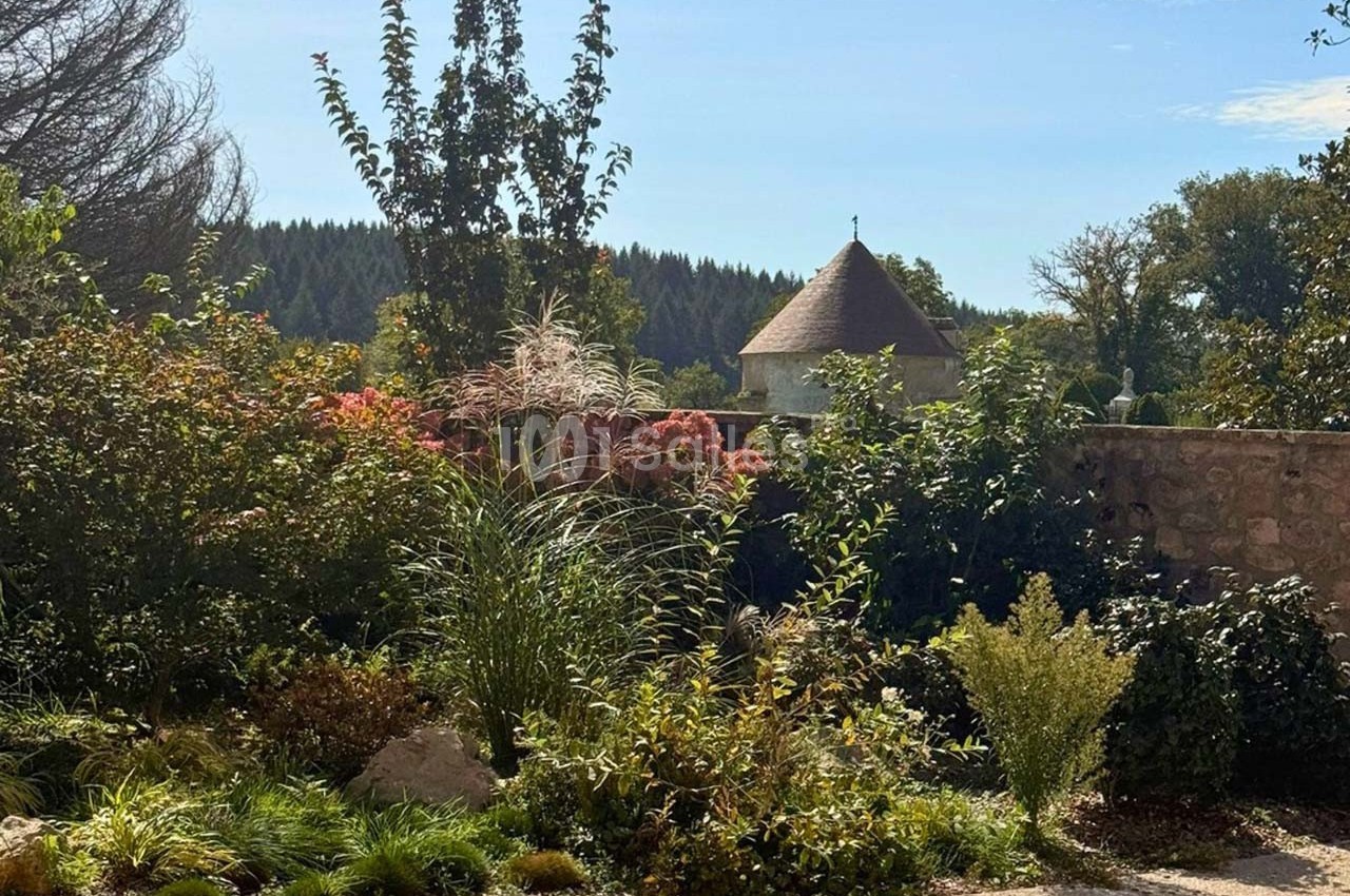 Jardin paysager avec arbustes, fleurs et arbres, devant un mur en pierre et une tour au toit conique sous un ciel bleu.
