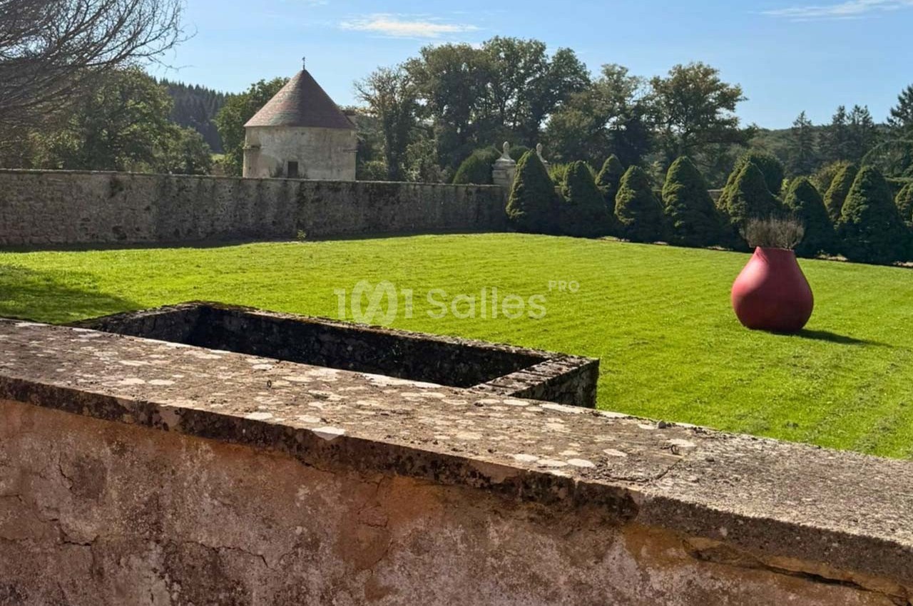 Vue d'un jardin avec pelouse, sculpture rouge en forme de vase, mur en pierre et tourelle en arrière-plan.