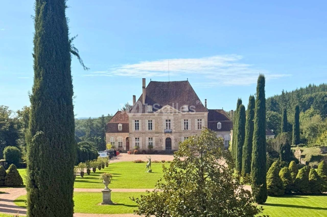 Manoir entouré de jardins verdoyants avec allées bordées de cyprès sous un ciel bleu clair.