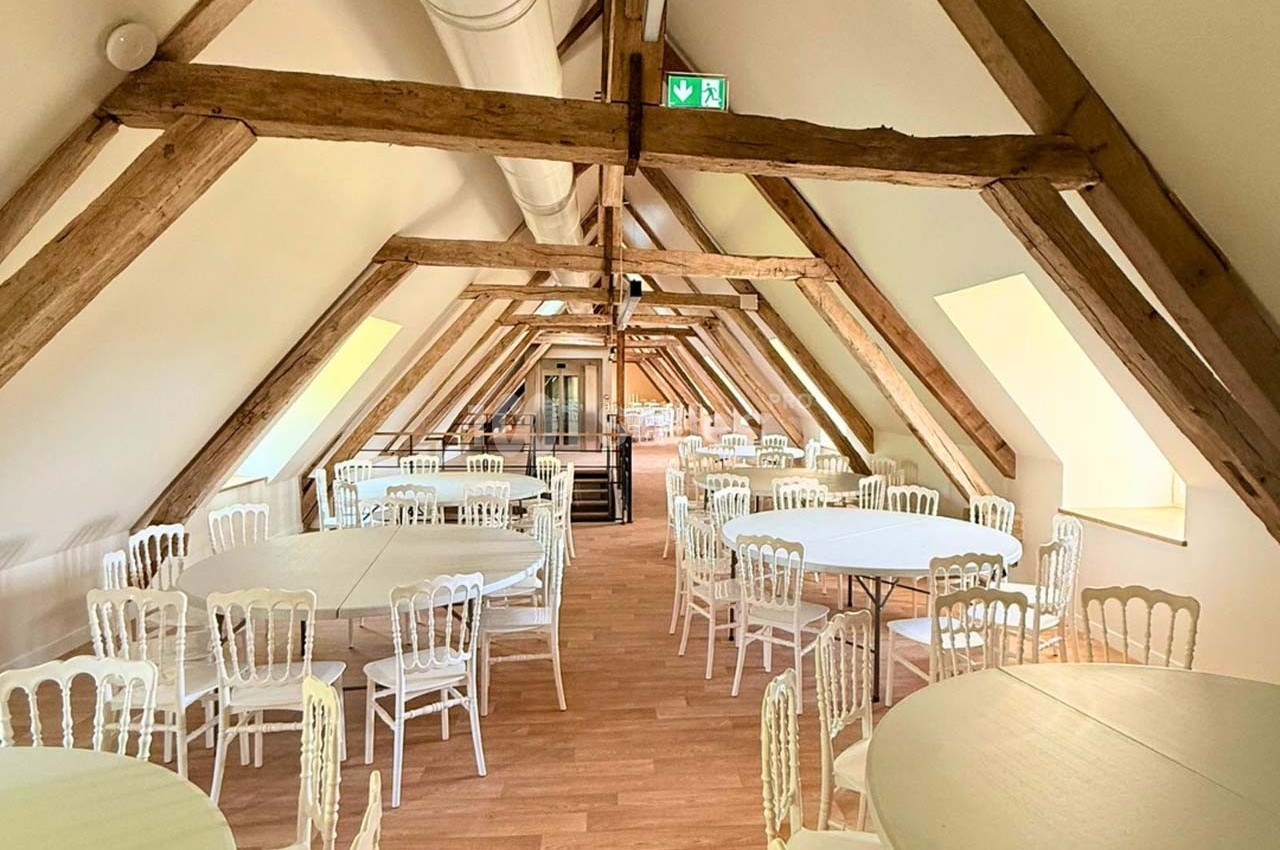 Salle lumineuse sous combles avec poutres apparentes, tables rondes et chaises blanches disposées pour un événement.
