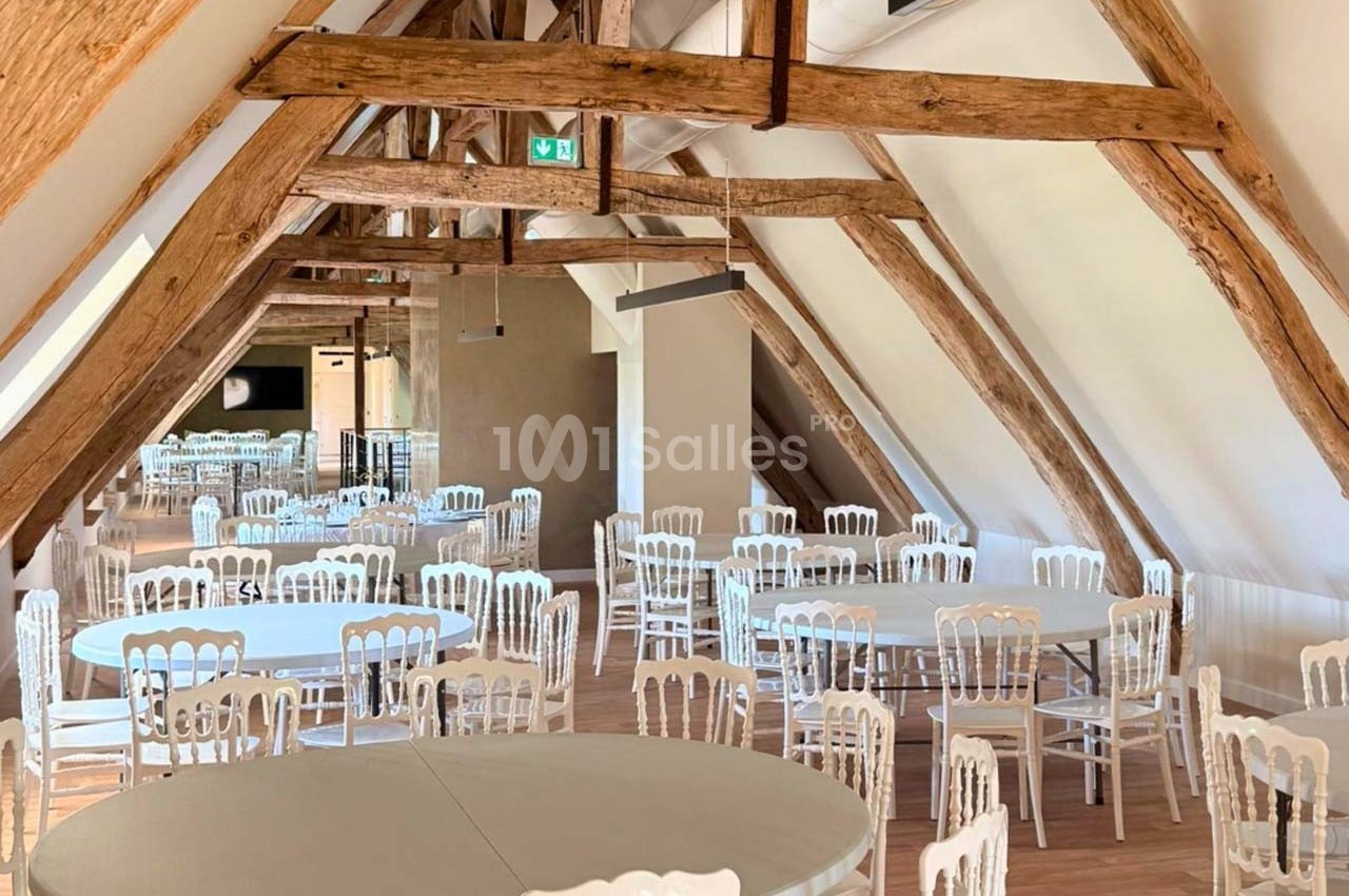 Salle lumineuse sous charpente en bois avec des tables rondes et des chaises blanches disposées pour un événement.