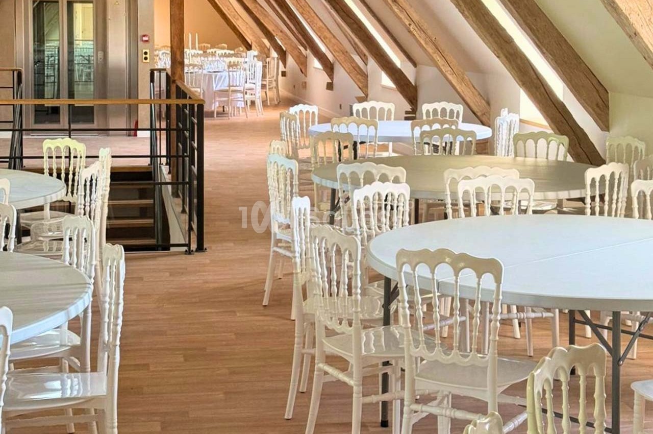 Salle lumineuse sous combles avec poutres apparentes, tables rondes et chaises blanches disposées pour un événement.