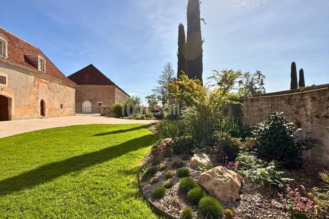 Jardin paysager avec pelouse, massifs de plantes et cyprès, bordé par des bâtiments en pierre sous un ciel ensoleillé.