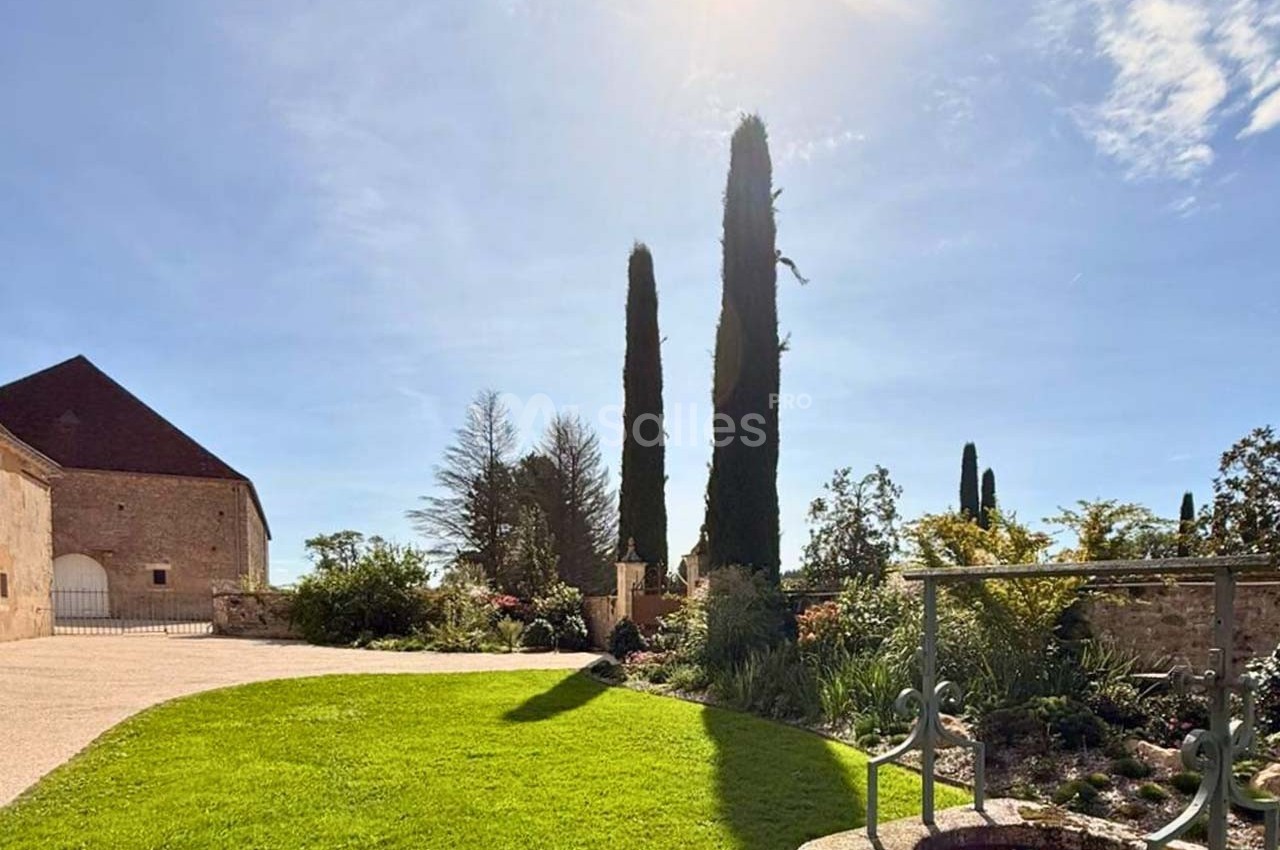 Jardin ensoleillé avec pelouse, puits en pierre, cyprès et bâtiment en pierre sous un ciel bleu dégagé.