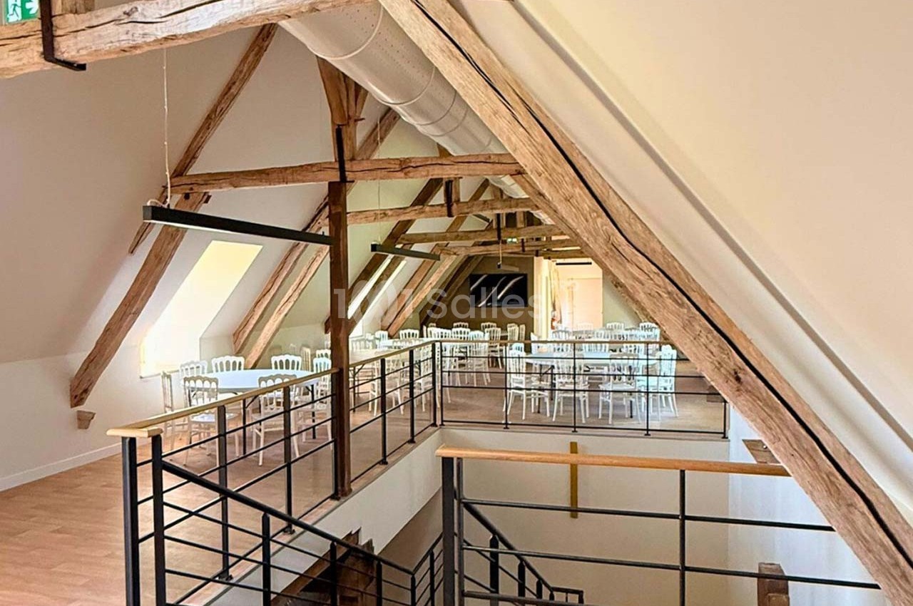 Salle lumineuse sous combles avec poutres apparentes, tables et chaises blanches, et escalier en métal et bois.