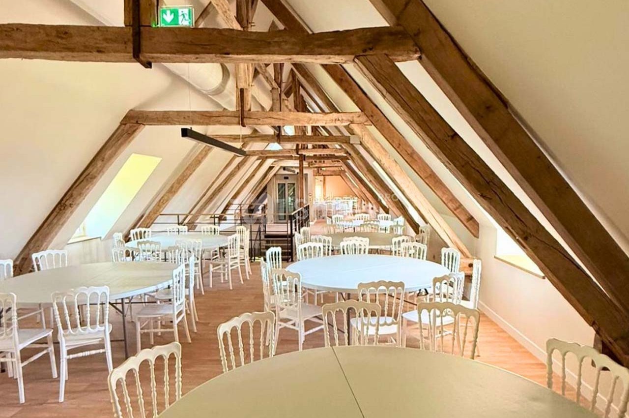 Salle sous combles avec poutres apparentes, tables rondes blanches et chaises alignées sur un sol en bois clair.