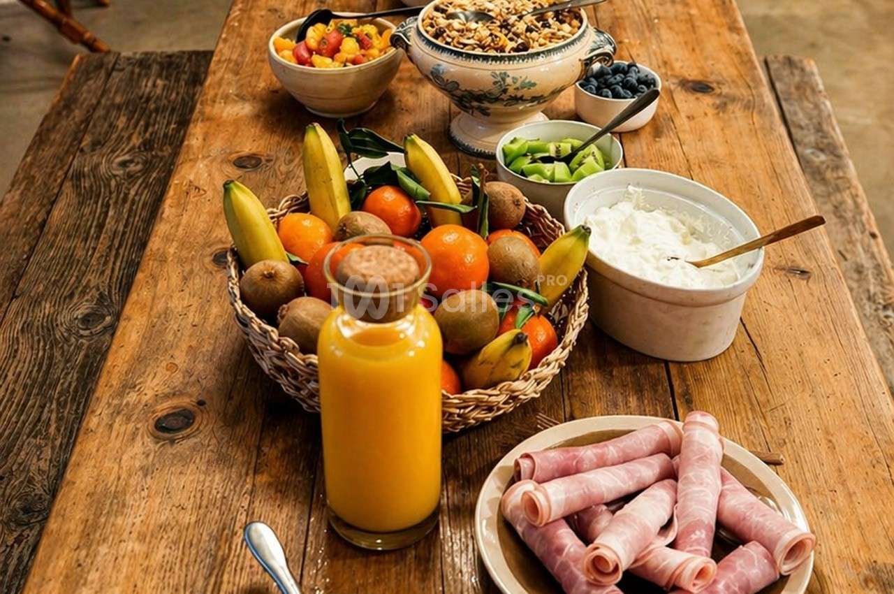 Petit-déjeuner sur une table en bois avec fruits, yaourt, céréales, jus d'orange et roulés de jambon.