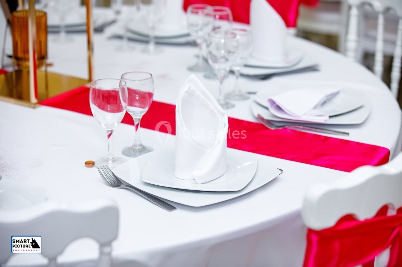 Table dressée avec une nappe blanche, serviettes pliées, verres, couverts et chemin de table rouge.