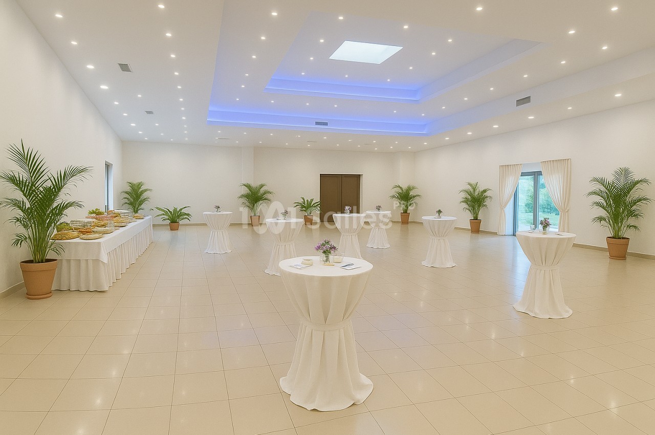 Salle lumineuse avec tables hautes drapées de blanc, plantes en pot et buffet disposé sur le côté.