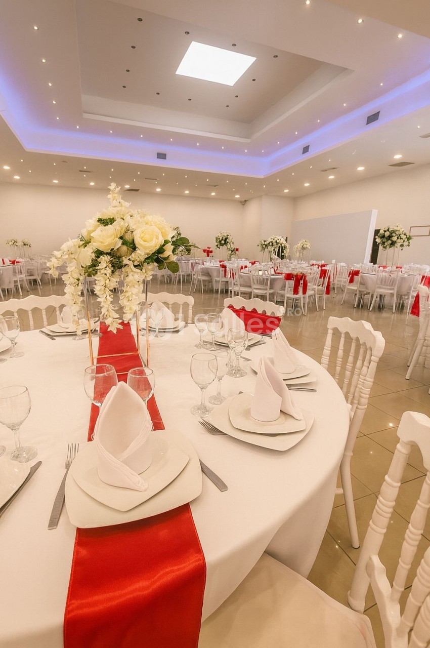 Salle de réception décorée avec des tables rondes, nappes blanches, chemins de table rouges et centres de table floraux.
