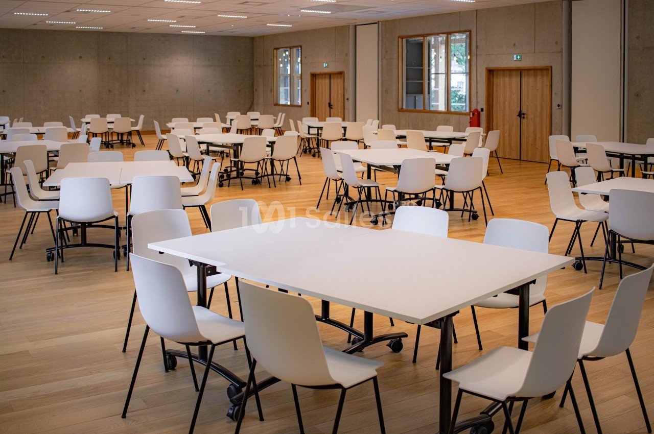Salle spacieuse avec des tables blanches et des chaises alignées sur un sol en bois clair, éclairée par des fenêtres.