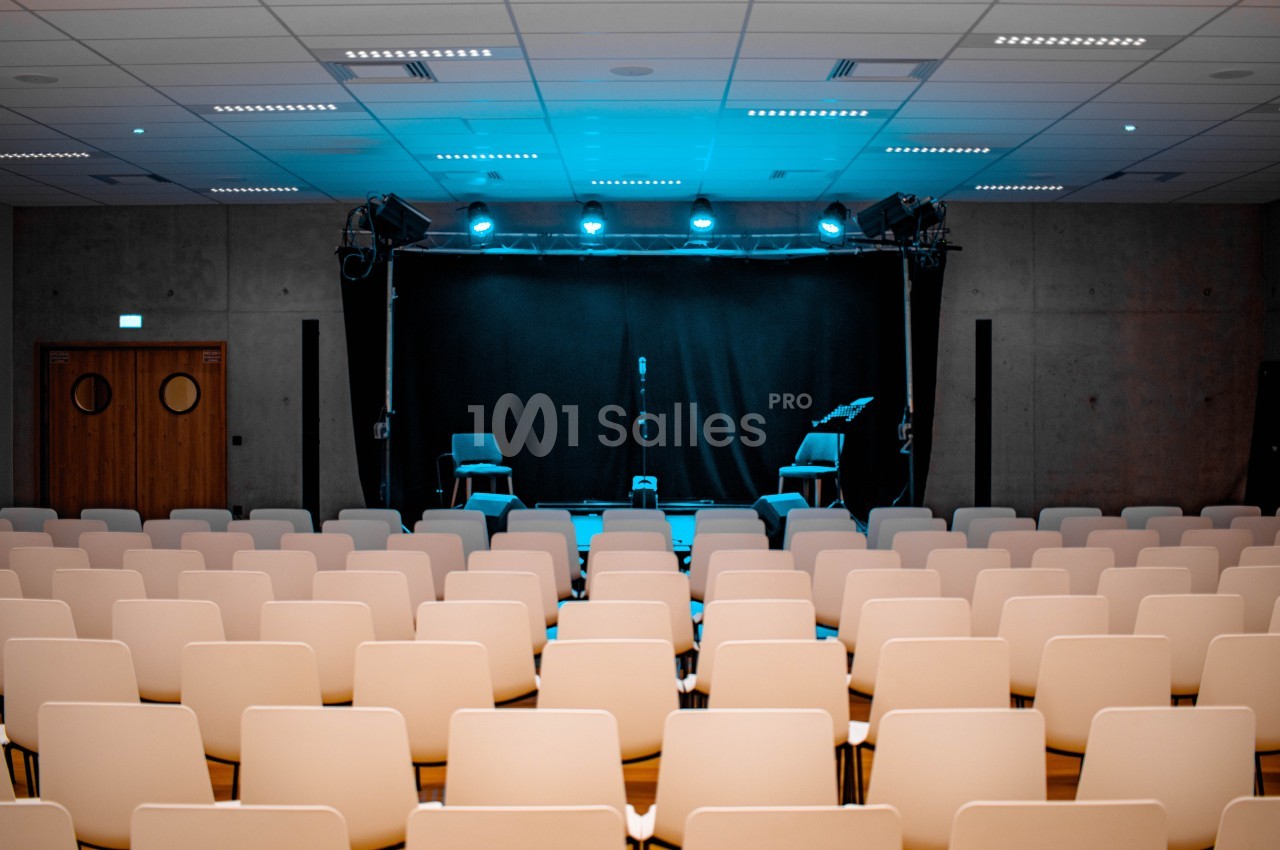 Salle de spectacle vide avec des rangées de chaises blanches face à une scène éclairée par des projecteurs bleus.