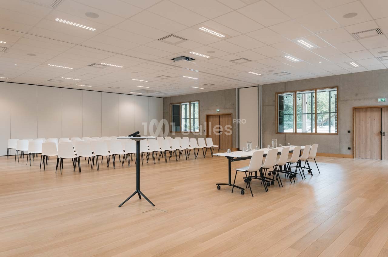 Salle de conférence lumineuse avec chaises blanches alignées, pupitre noir et table équipée de matériel.