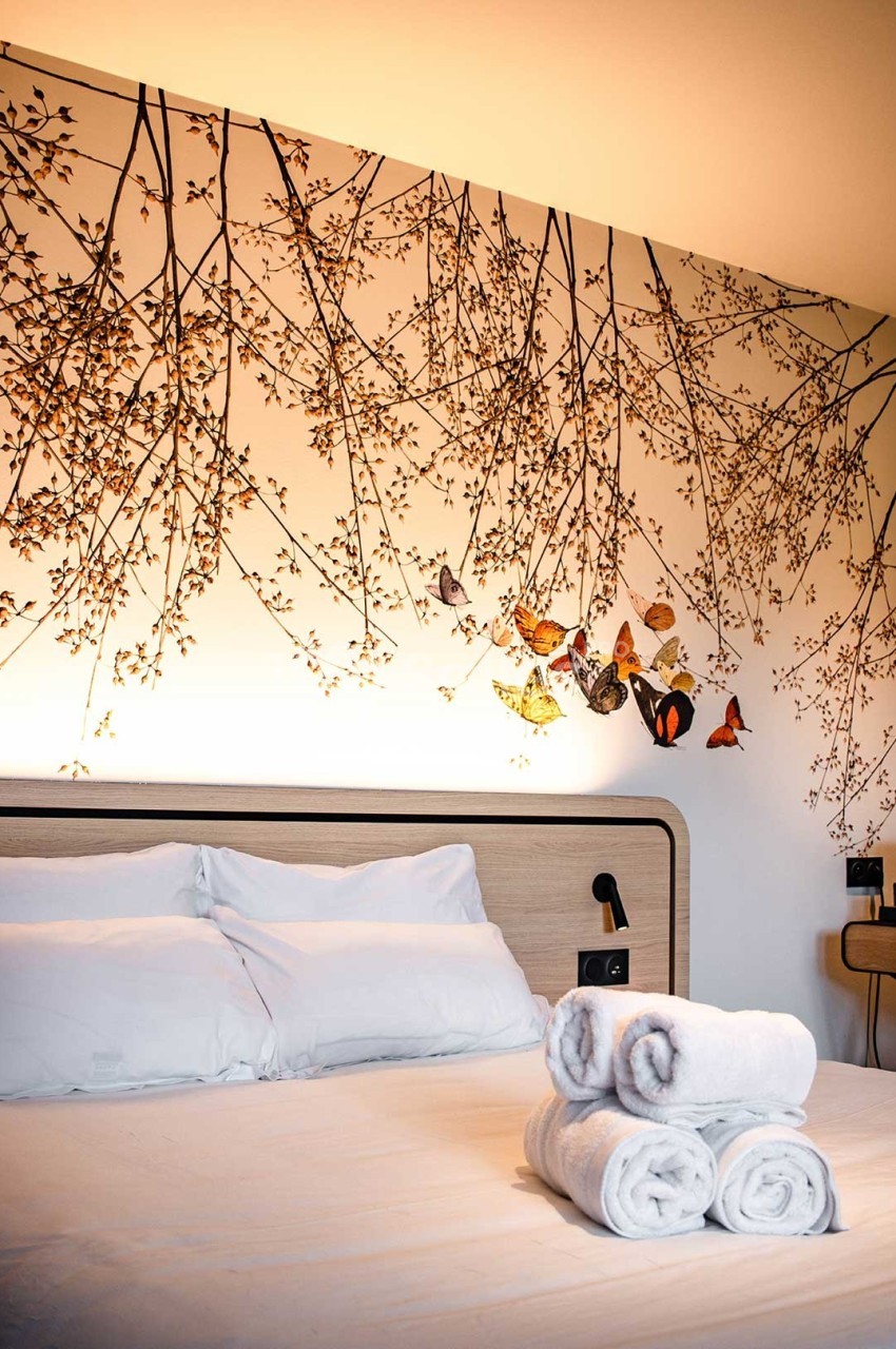 Lit avec des draps blancs, serviettes pliées au premier plan et mur décoré de branches et papillons colorés.
