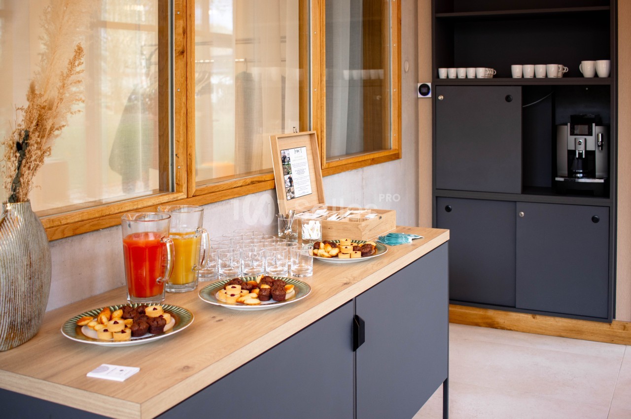 Buffet avec jus de fruits, biscuits et verres sur un meuble, près d'une machine à café et d'armoires grises.