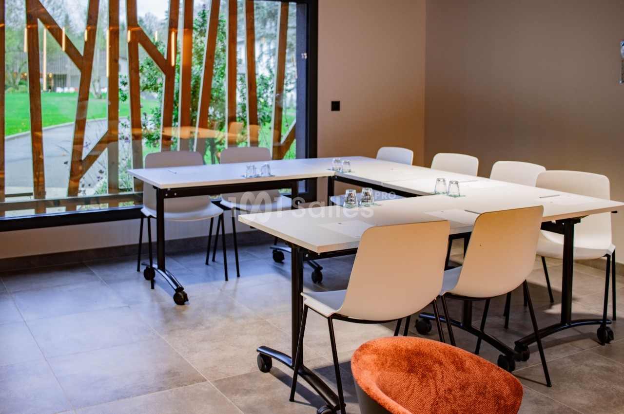 Salle de réunion lumineuse avec des tables modulables, des chaises blanches et une grande baie vitrée donnant sur un jardin.