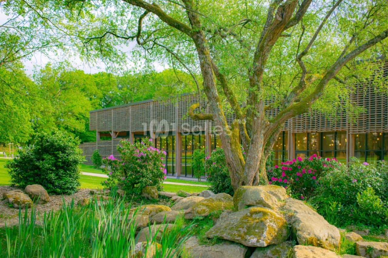 Bâtiment moderne entouré de verdure, avec un arbre au premier plan et des arbustes fleuris.
