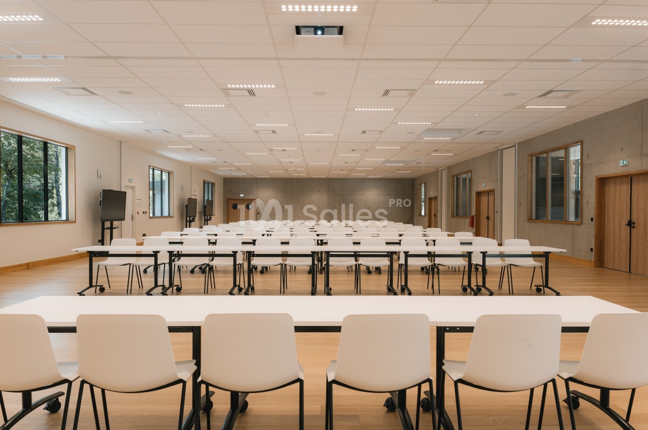 Salle de conférence vide avec des rangées de tables et de chaises blanches, éclairée par des lumières au plafond.