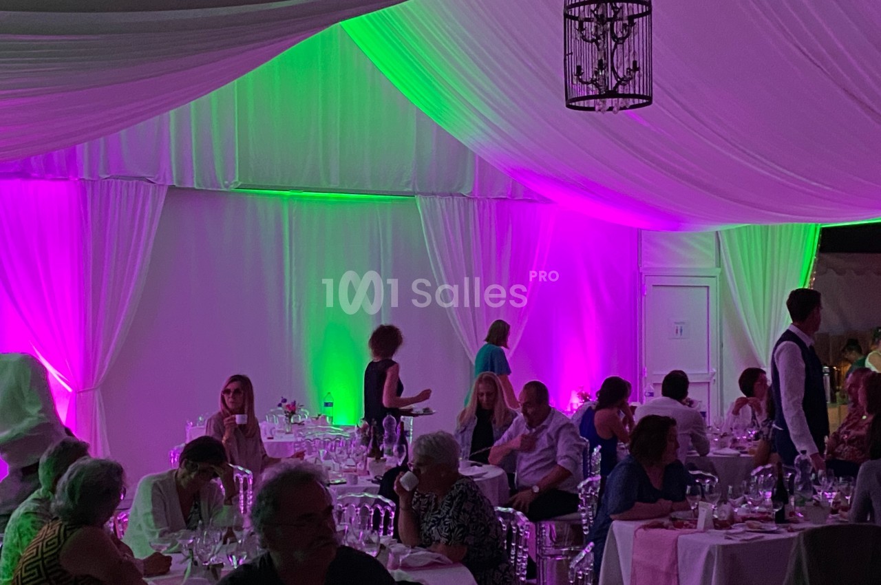 Salle de réception avec des invités dînant sous une tente éclairée par des lumières violettes et vertes.