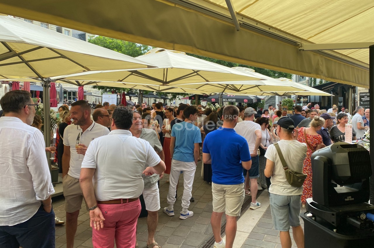 Groupe de personnes rassemblées sous des parasols dans une rue animée, discutant et partageant un moment convivial.