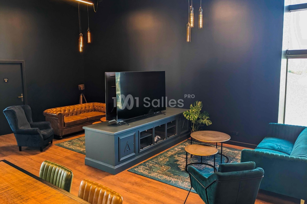 Salon moderne avec canapé en cuir, fauteuils, tapis coloré, meuble TV et éclairage suspendu dans une pièce sombre.