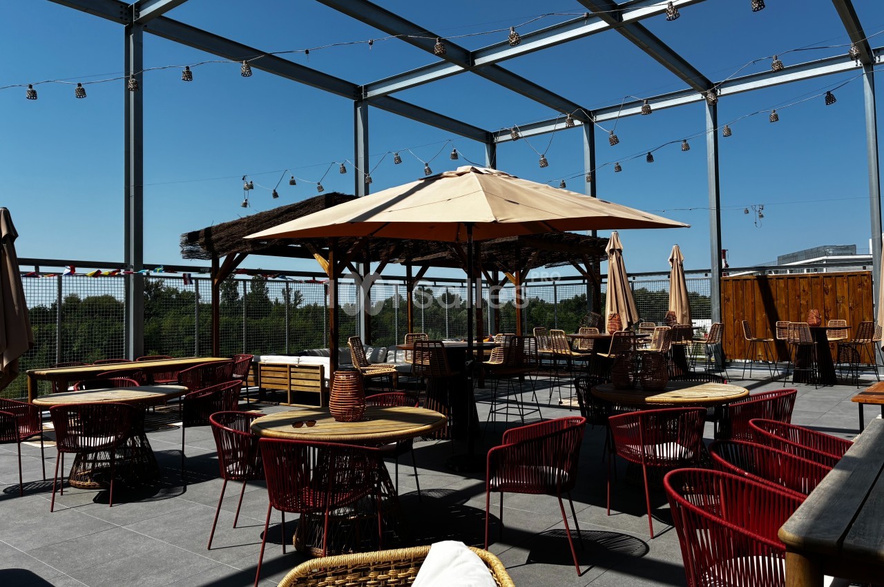 Terrasse ensoleillée avec tables, chaises rouges et parasols, entourée de guirlandes lumineuses et d'une structure…