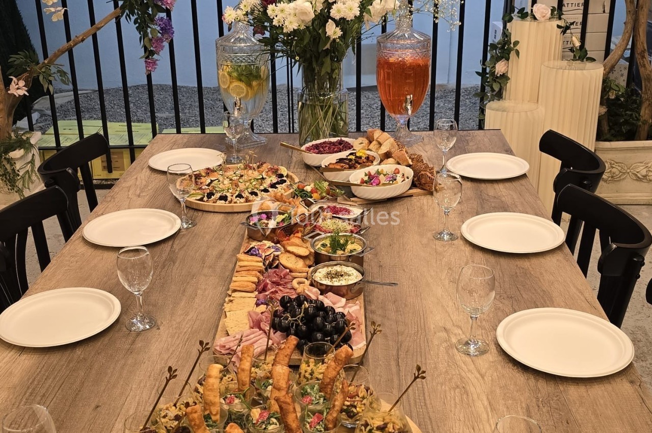 Table en bois dressée avec des plats variés, verrines, charcuteries et boissons, entourée de chaises et décorée de fleurs.