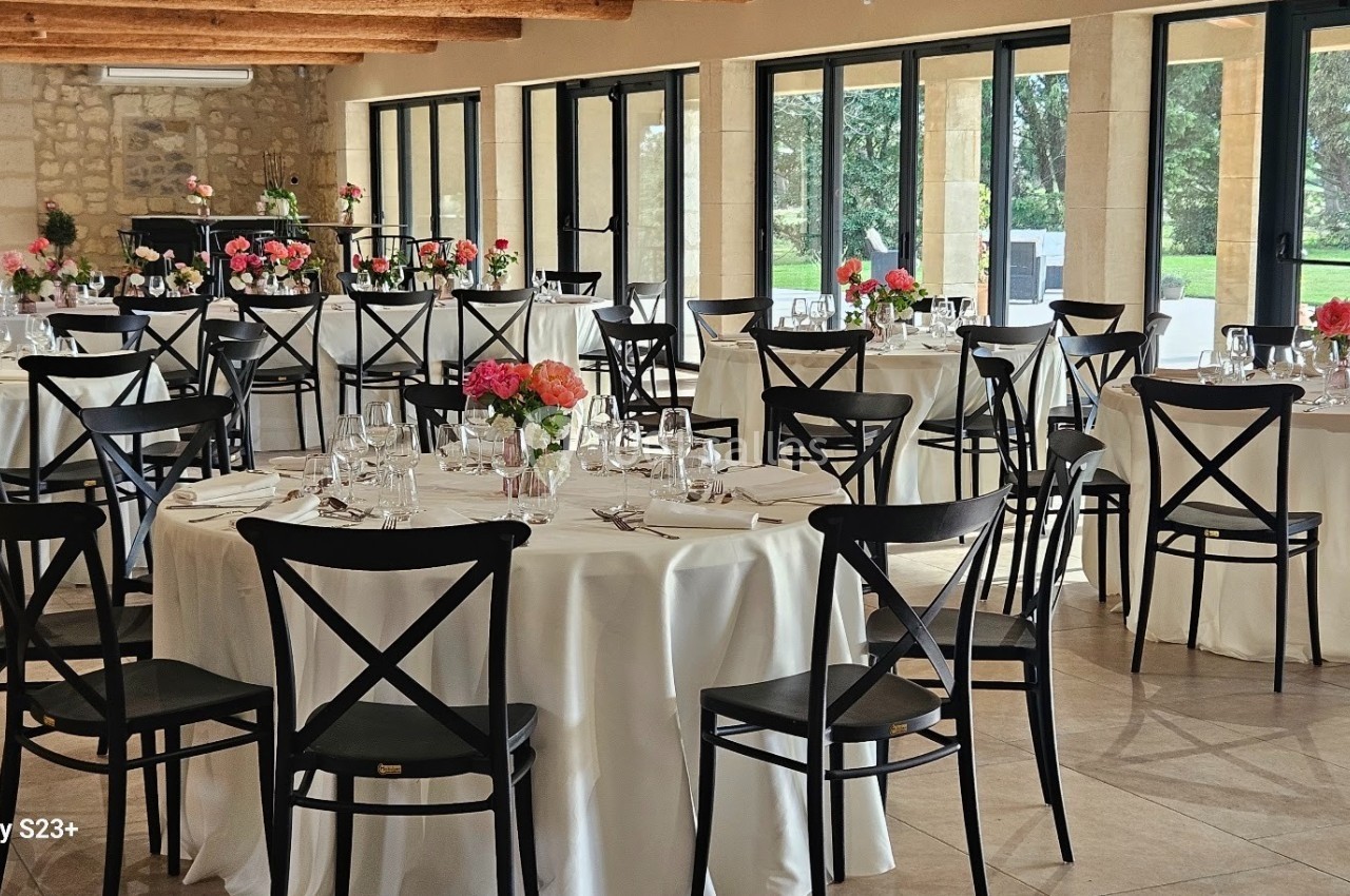 Salle de réception lumineuse avec tables rondes dressées, nappes blanches et chaises noires, décorées de fleurs colorées.