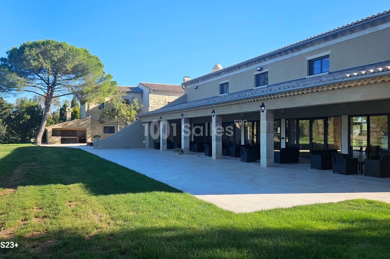 Grande maison moderne avec terrasse couverte, mobilier extérieur et jardin arboré sous un ciel dégagé.