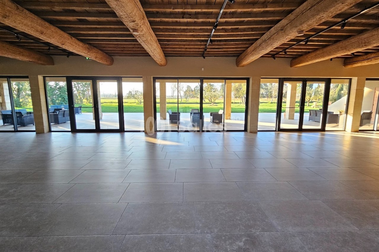 Salle spacieuse avec plafond en poutres apparentes, grandes baies vitrées donnant sur une terrasse et un espace vert.