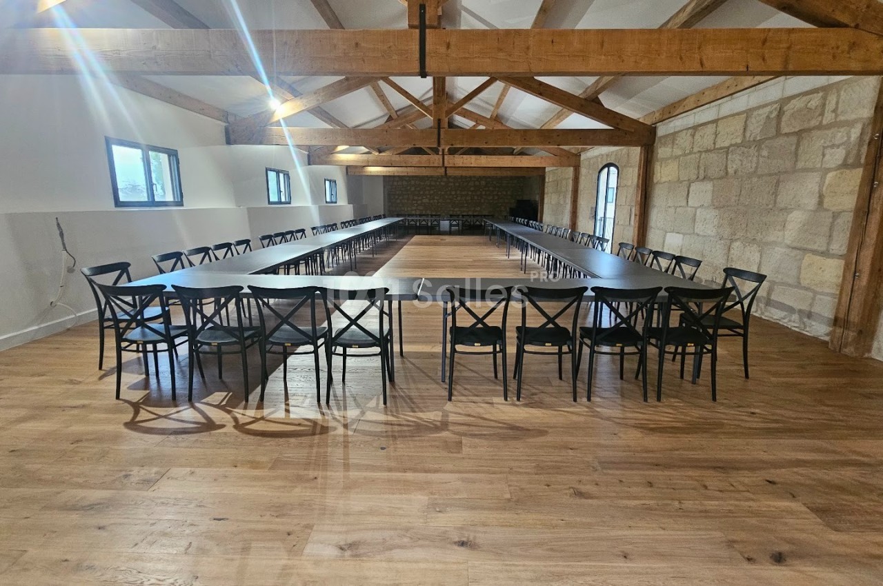 Salle lumineuse avec poutres apparentes, tables disposées en U et chaises noires sur un sol en parquet.