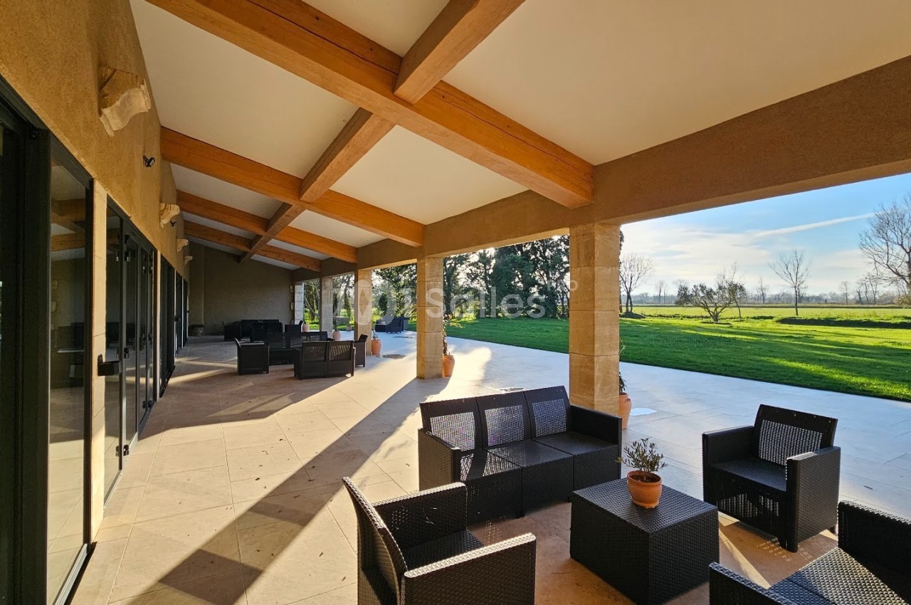 Terrasse couverte avec mobilier en rotin, donnant sur un jardin verdoyant et une grande pelouse ensoleillée.