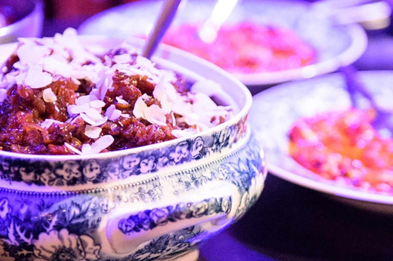 Plat en céramique décorée contenant un mets garni d'amandes effilées, entouré d'assiettes colorées floues.