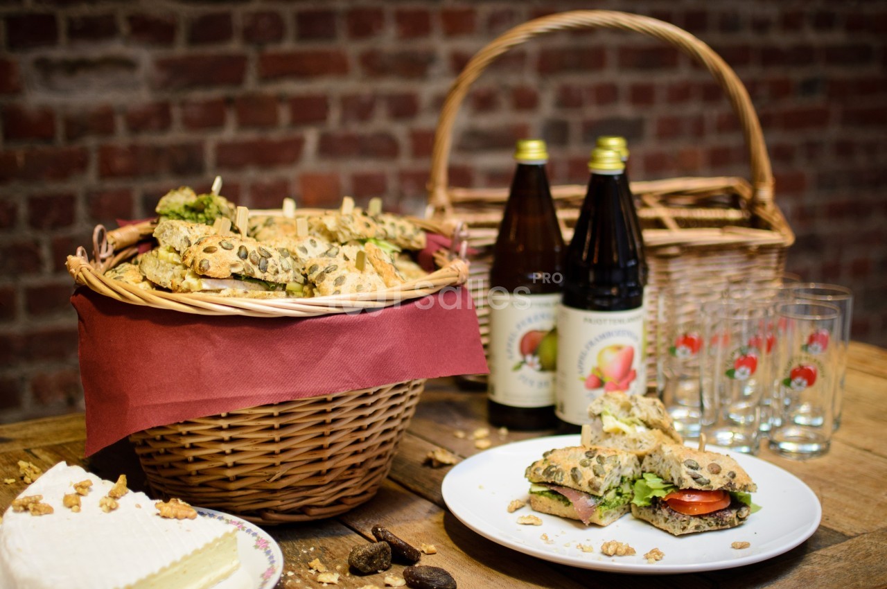 Panier de sandwichs garnis, bouteilles de jus de pomme, verres et assiette de fromage sur une table en bois.