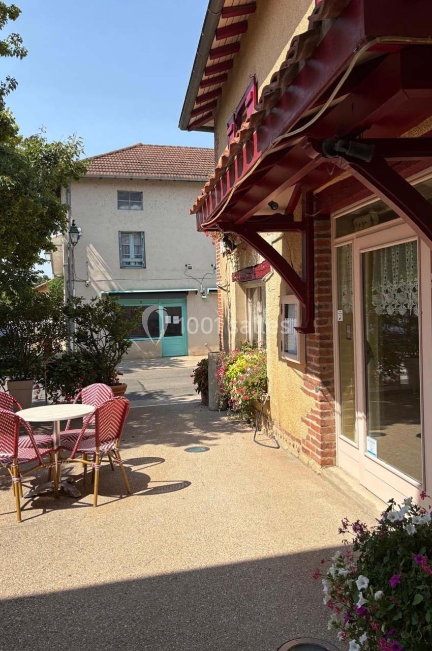 Terrasse avec table et chaises rouges devant un bâtiment, entourée de fleurs, sous un ciel dégagé.