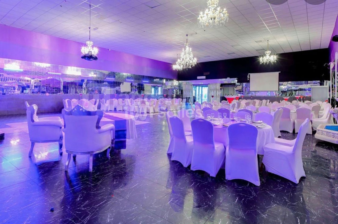 Salle de réception élégante avec tables rondes dressées, chaises blanches et lustres suspendus.