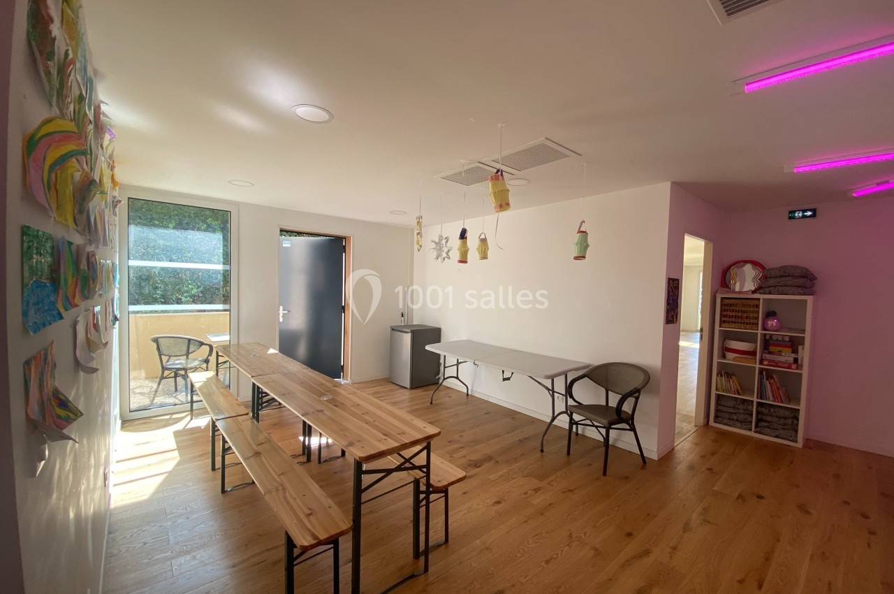 Salle lumineuse avec tables en bois, chaises, décorations suspendues et dessins d'enfants accrochés au mur.