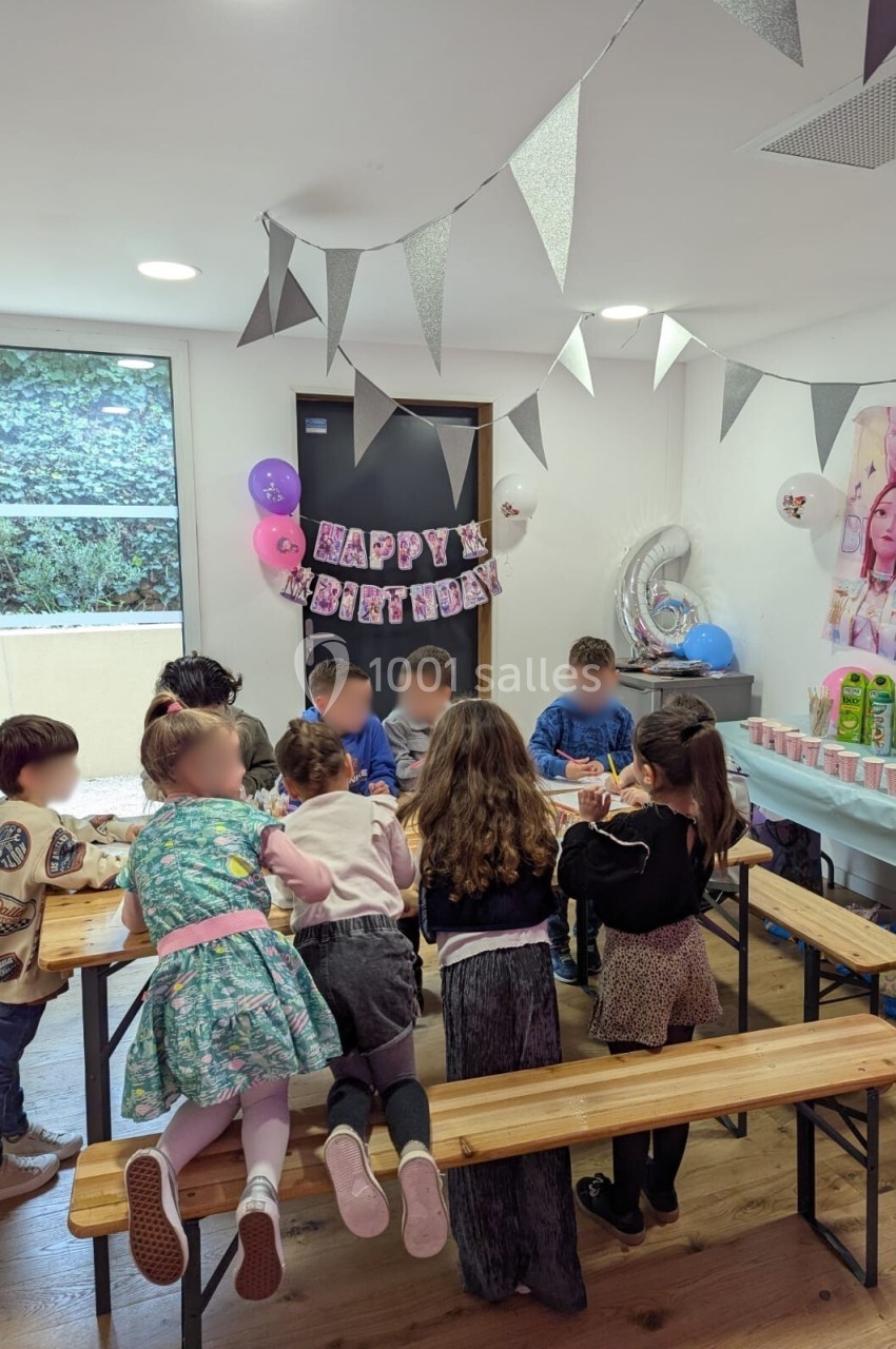 Des enfants assis autour de tables en bois décorées, célébrant un anniversaire dans une pièce ornée de ballons et guirlandes.