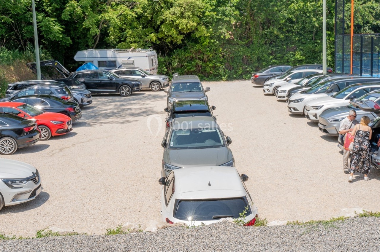 Parking extérieur avec plusieurs voitures stationnées sur un sol gravillonné, entouré de végétation et d'un terrain de sport.