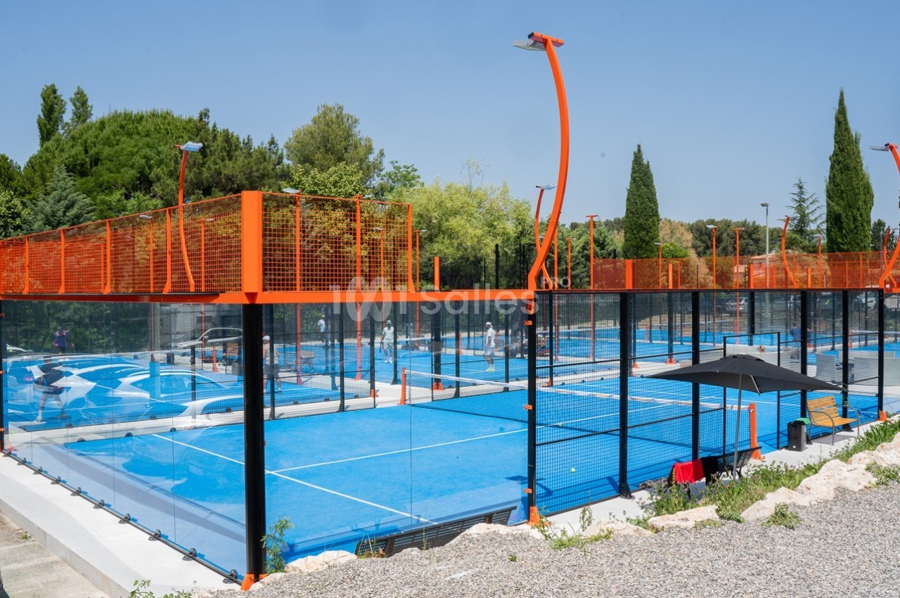 Terrains de padel extérieurs avec revêtement bleu, entourés de grillages orange, situés dans un espace arboré.