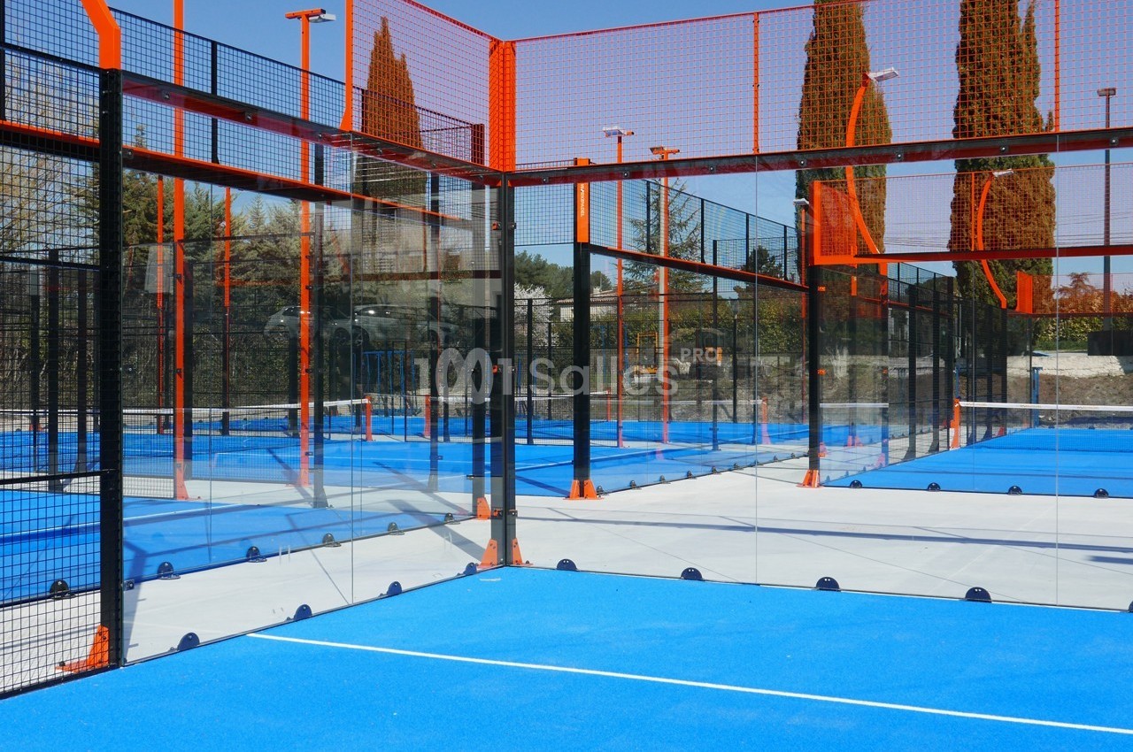 Terrains de padel extérieurs avec sol bleu, entourés de grillages et de parois vitrées, sous un ciel dégagé.