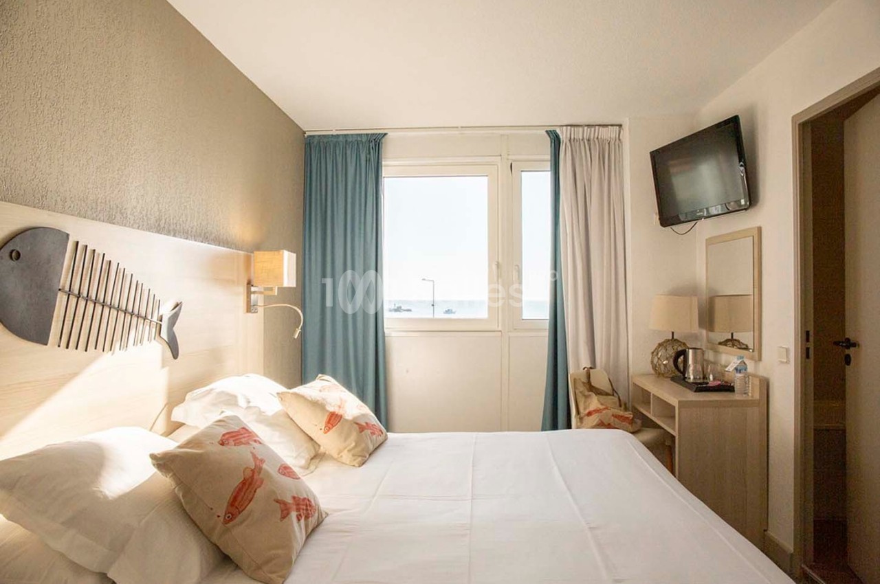 Chambre lumineuse avec lit double, décoration marine, fenêtre donnant sur une vue extérieure et télévision murale.